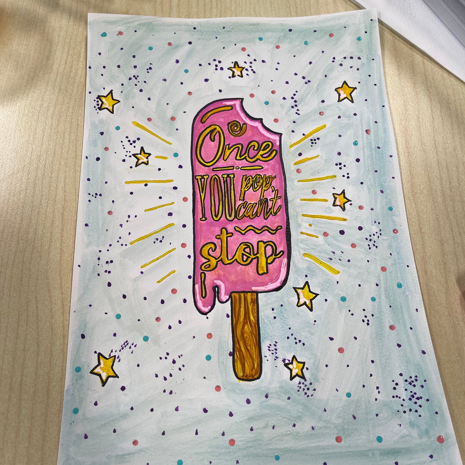 Colorful drawing of a popsicle with text on a light blue background, placed on a wooden surface.